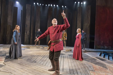 Lear in red tunk and brown pants pointing up remonstratively as Goneril in blue cloak and Regan in red dress look on in irritation
